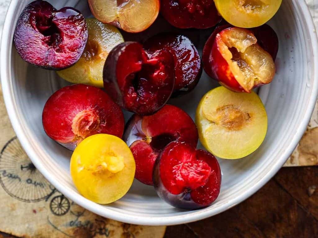 A white bowl filled with halved red and yellow plums, showing their juicy flesh and glossy skins, sits on a table with part of a vintage-style paper visible underneath.
